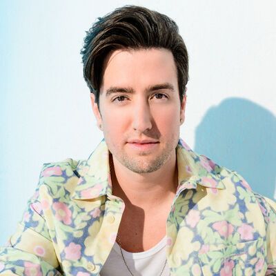 A person with dark hair styled upward, wearing a white t-shirt and a floral yellow shirt against a light blue background.