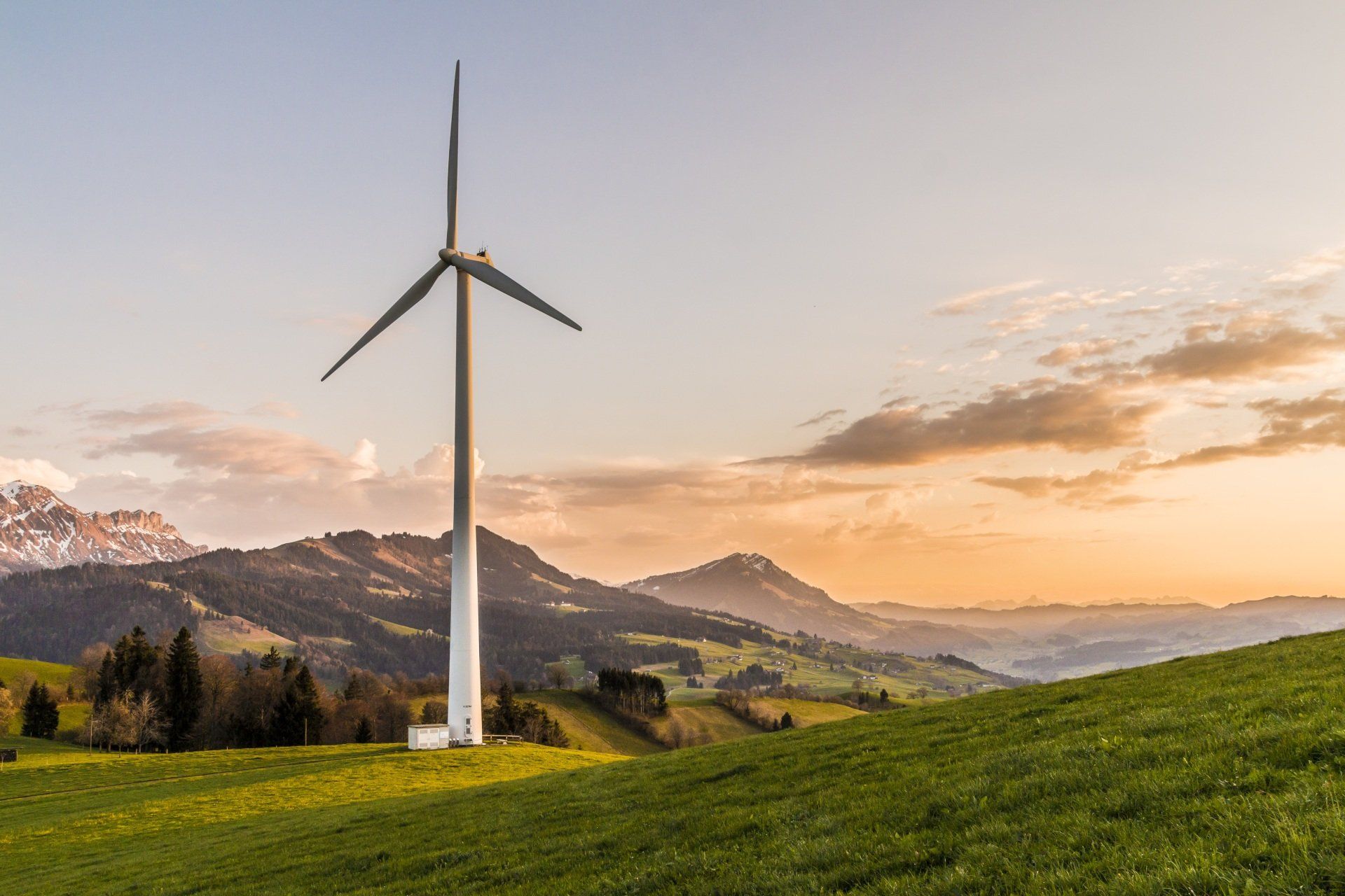 A single wind turbine stands in a lush green field before rolling mountains under a golden, sunset-lit sky.