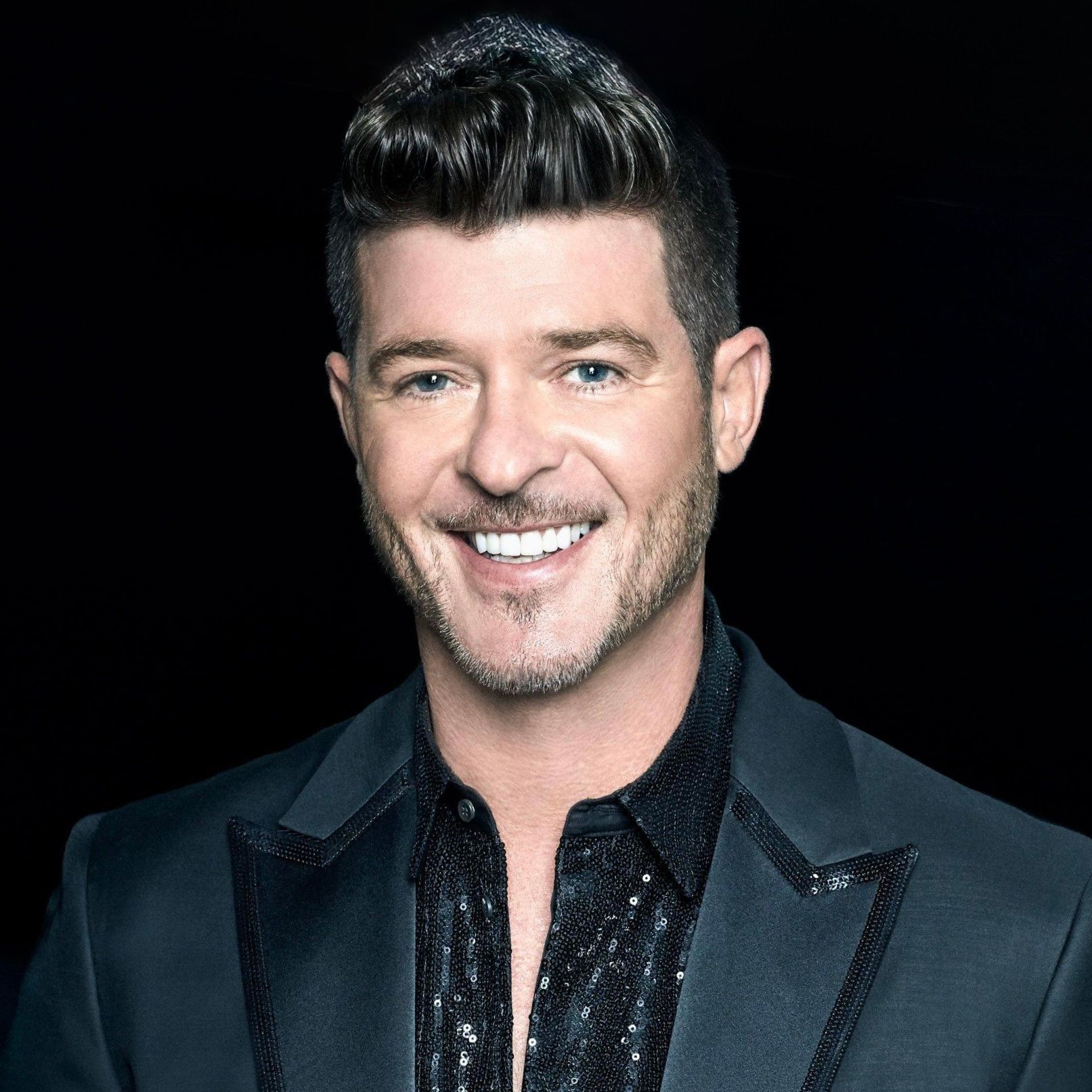 Robin Thicke wearing a dark, sequined shirt and a matching blazer against a black background.