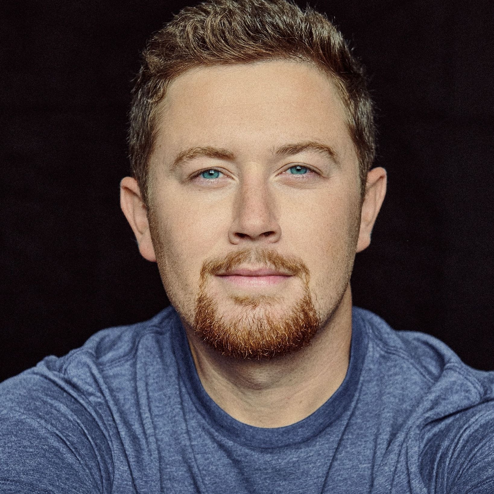 A close-up portrait of a person with short brown hair and a goatee, wearing a blue t-shirt against a black background.