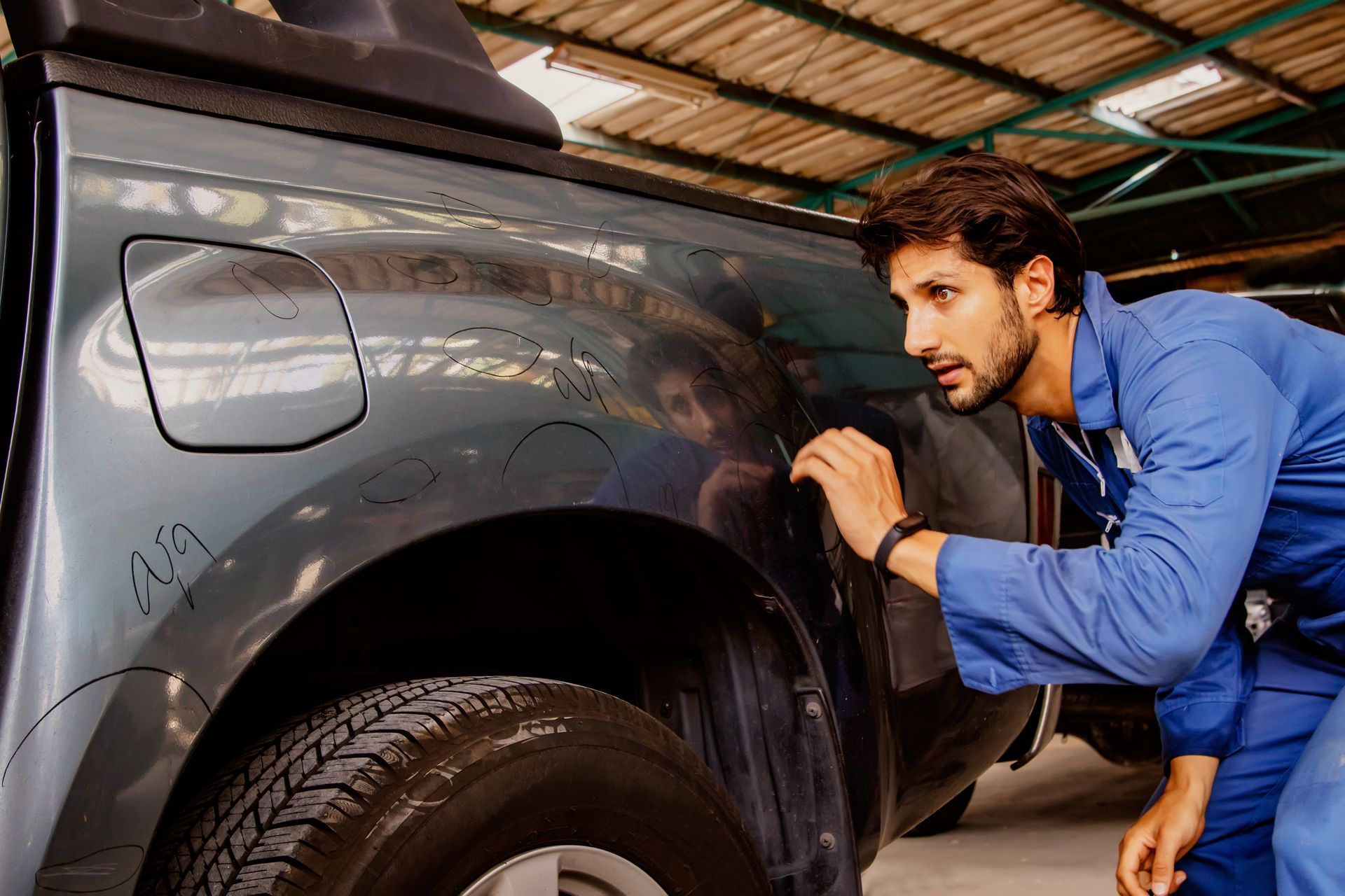 Mechanic in blue overalls inspecting a gray car’s rear wheel area in a garage