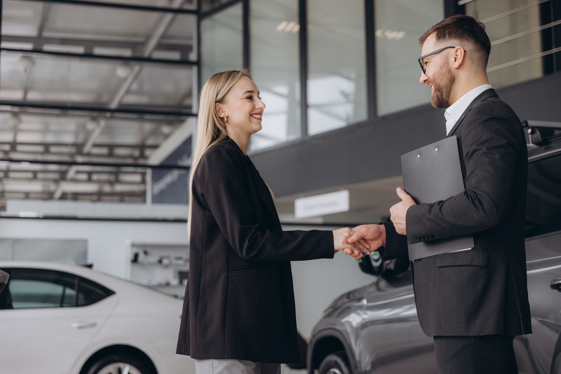 Two professionals shake hands in a car showroom beside a white car.