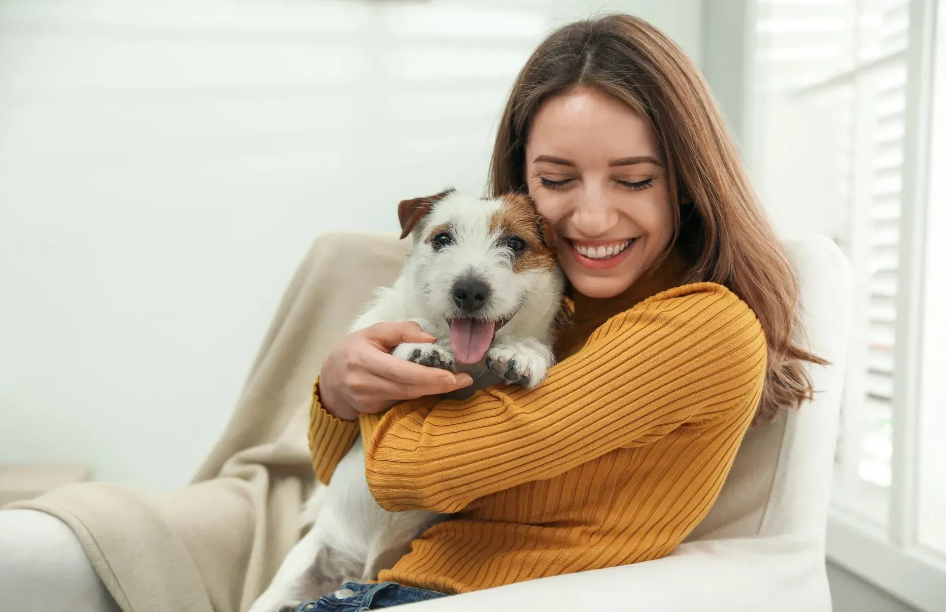 A woman in a yellow sweater sits on a chair, smiling with her eyes closed, while holding a small white and brown dog that is panting.