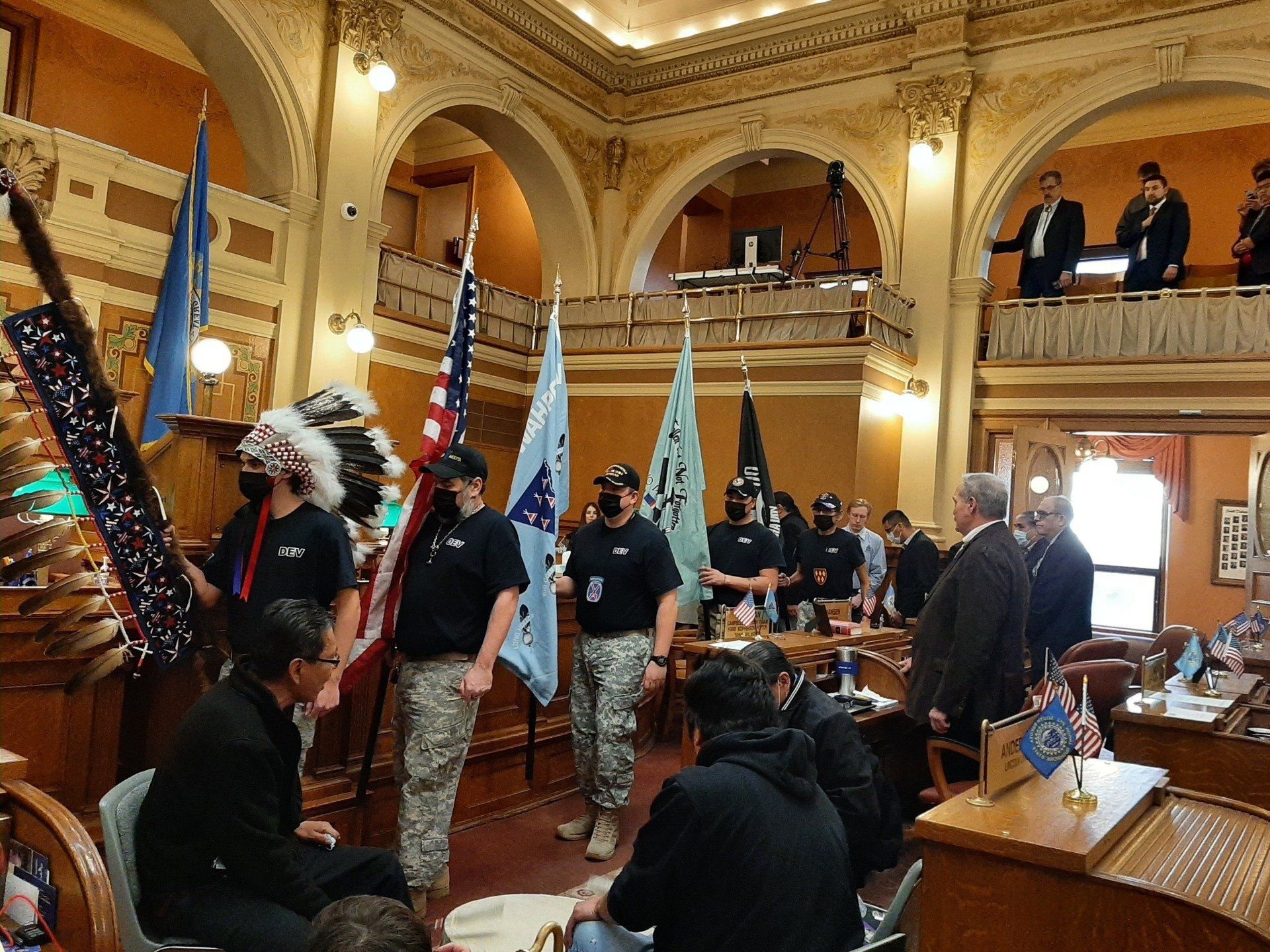 Group with flags and headdresses in a legislative chamber. People in suits watch from a balcony.