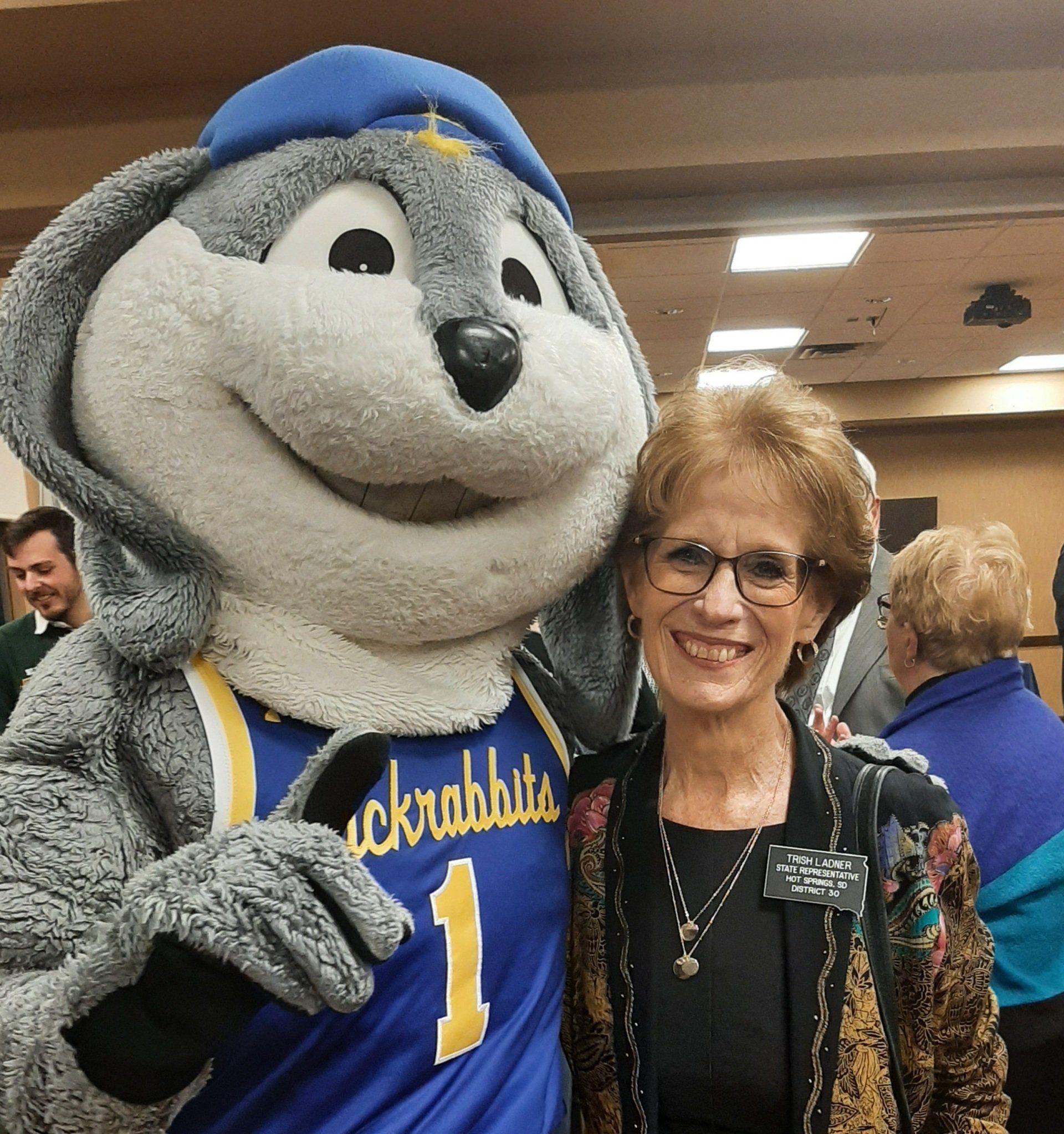 Woman smiles next to a Jackrabbit mascot wearing a basketball jersey; indoors with other people.