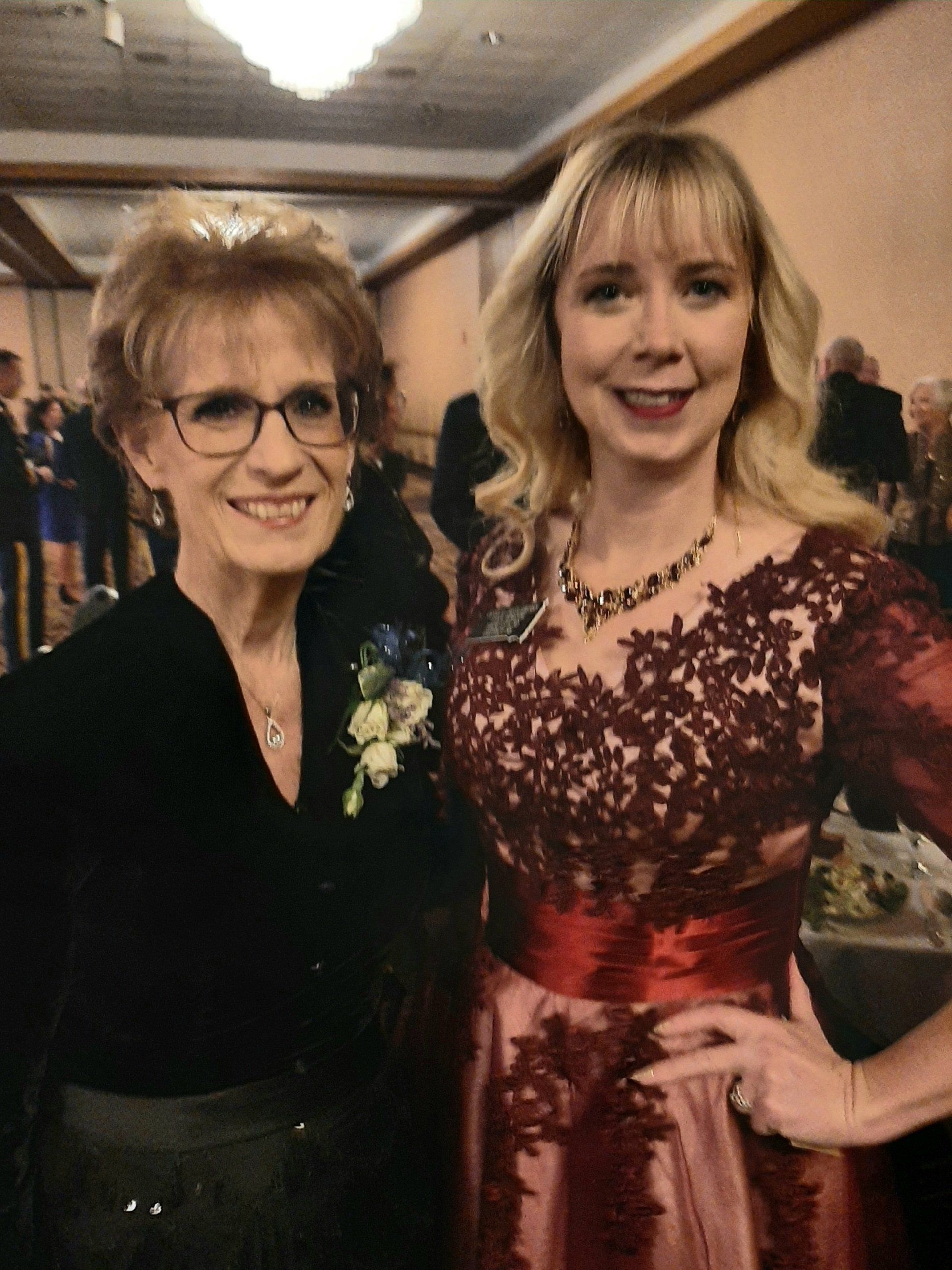 Woman with glasses smiles with a woman in a red lace dress at a formal event.