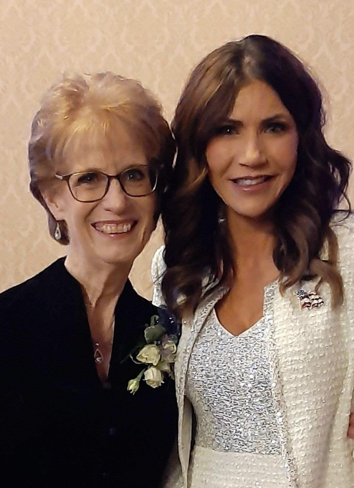 Two women pose together; one smiles, wearing glasses and a black top. The other smiles, wearing a cream blazer.