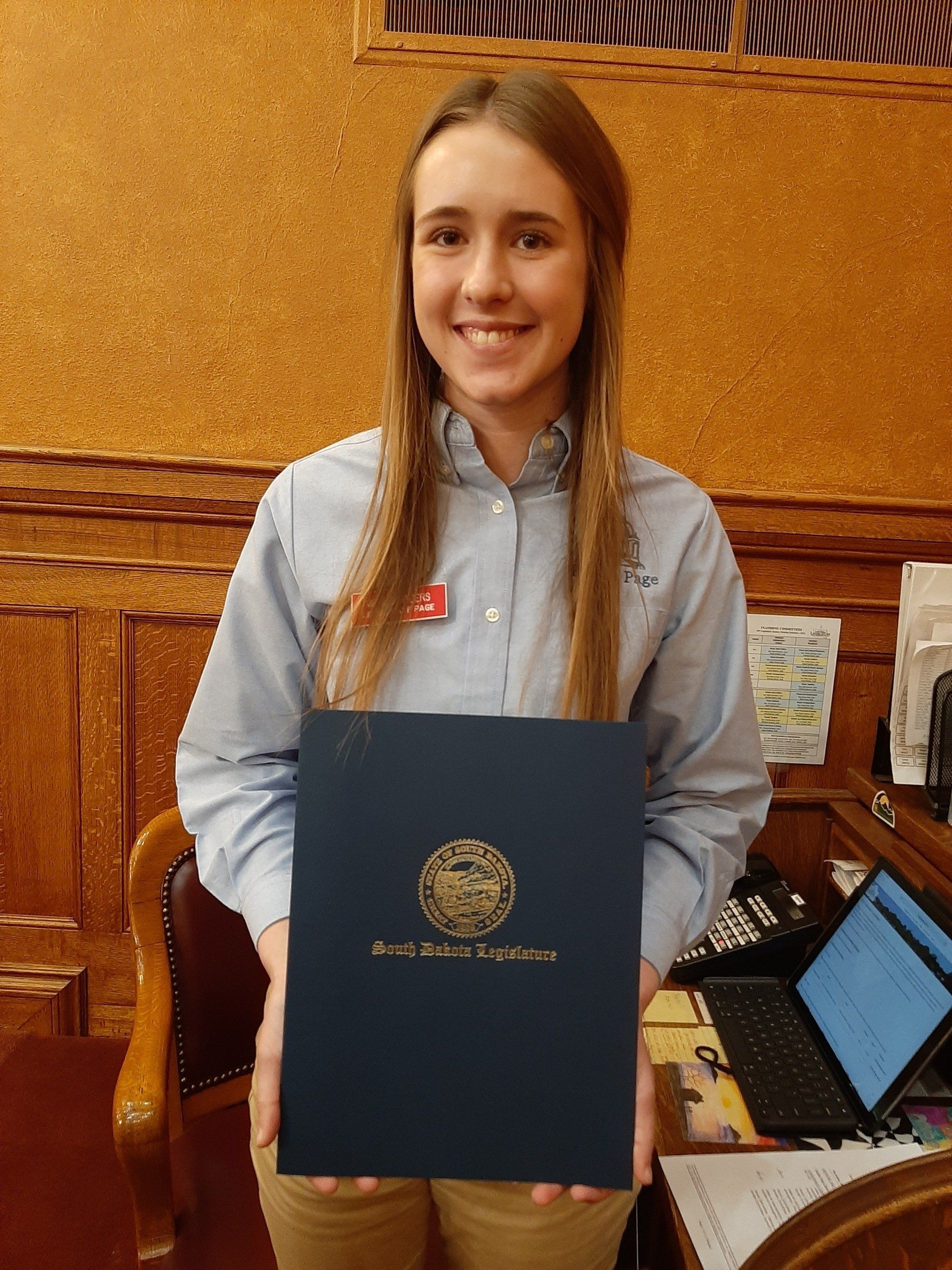 Woman smiling, holding a blue document with gold seal in an office setting.