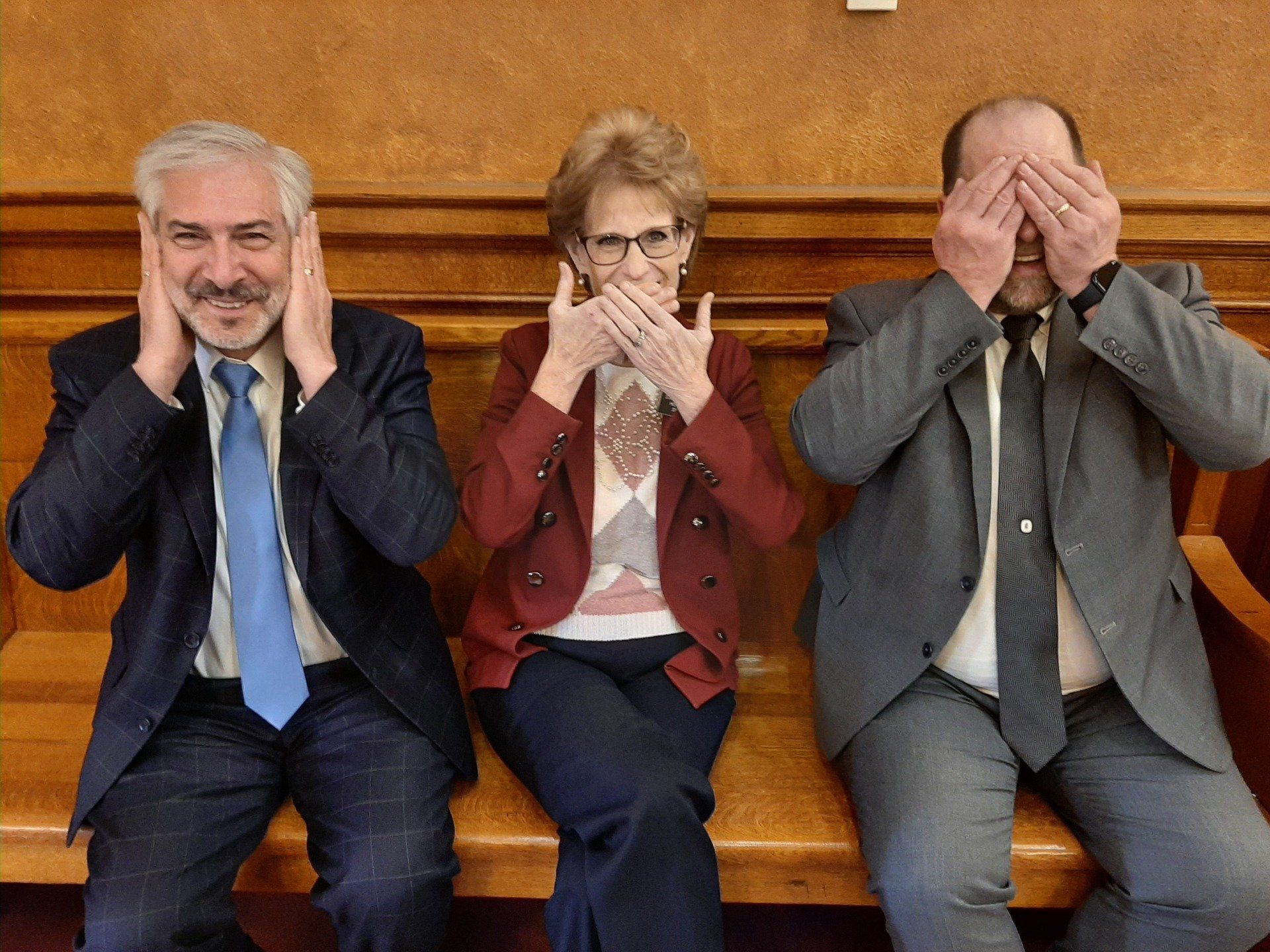 Three people seated, each covering an area of their face: ears, mouth, and eyes, inside a wood-paneled room.