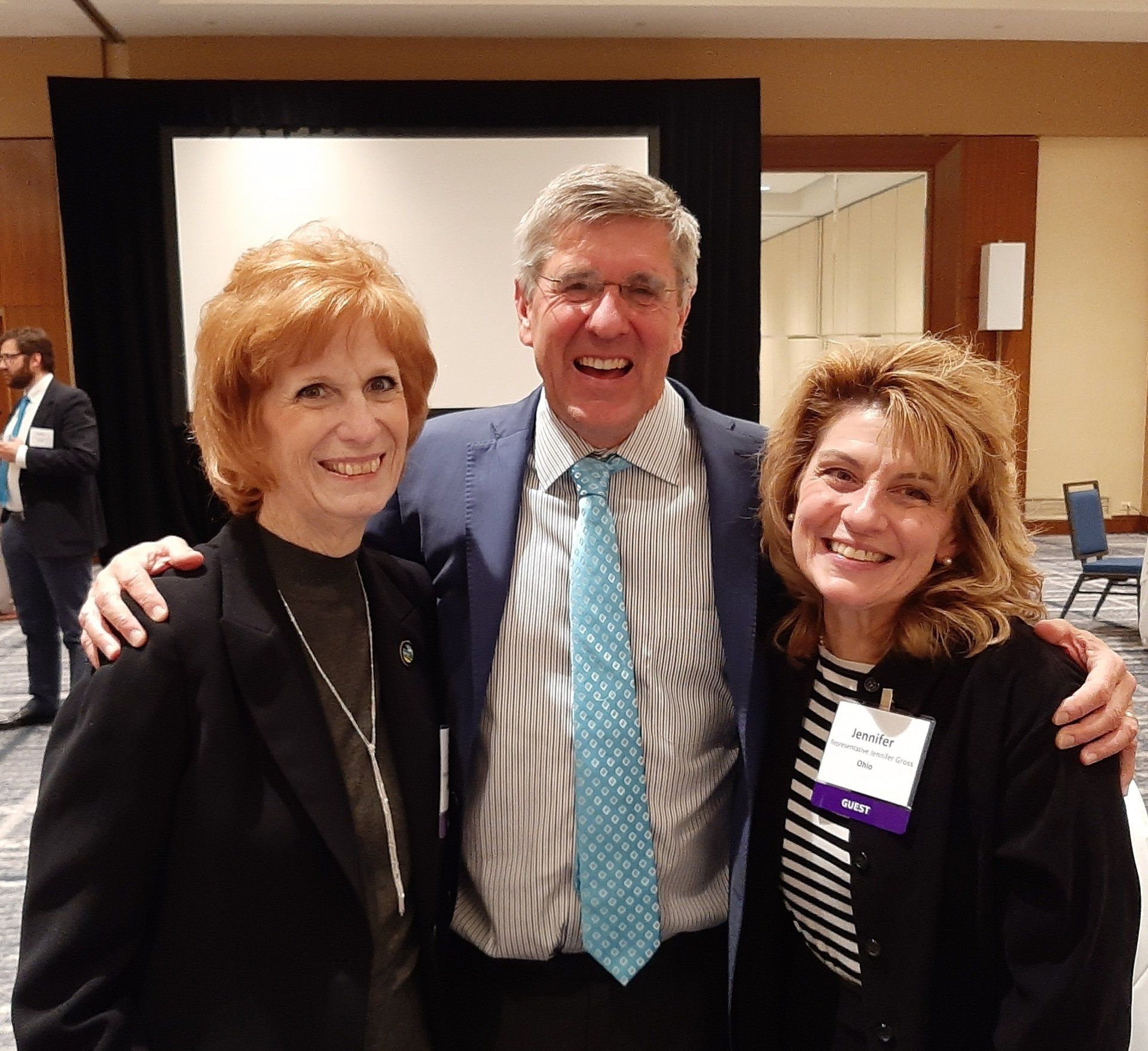 Three people smiling, posing for photo in front of a screen and a conference setting.