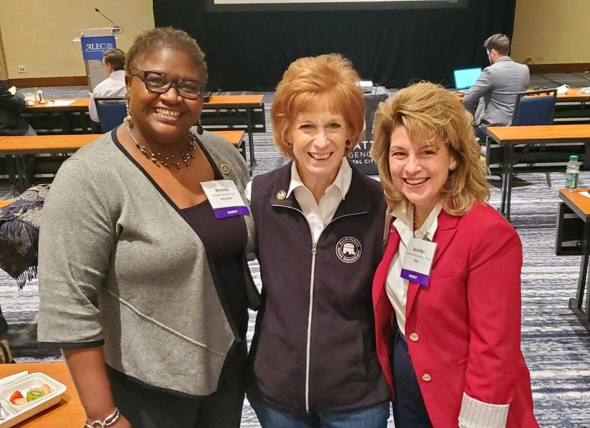 Three women smiling, posing for a photo in a conference room. One in gray, one in a vest, one in a red blazer.