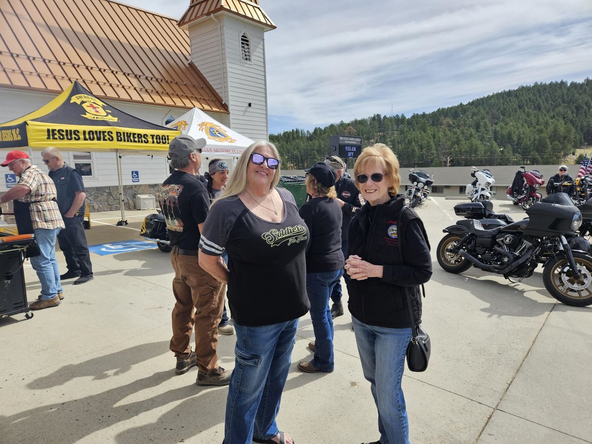 Two people standing in front of parked motorcycles at a lakeside event, with a sign and tower behind them