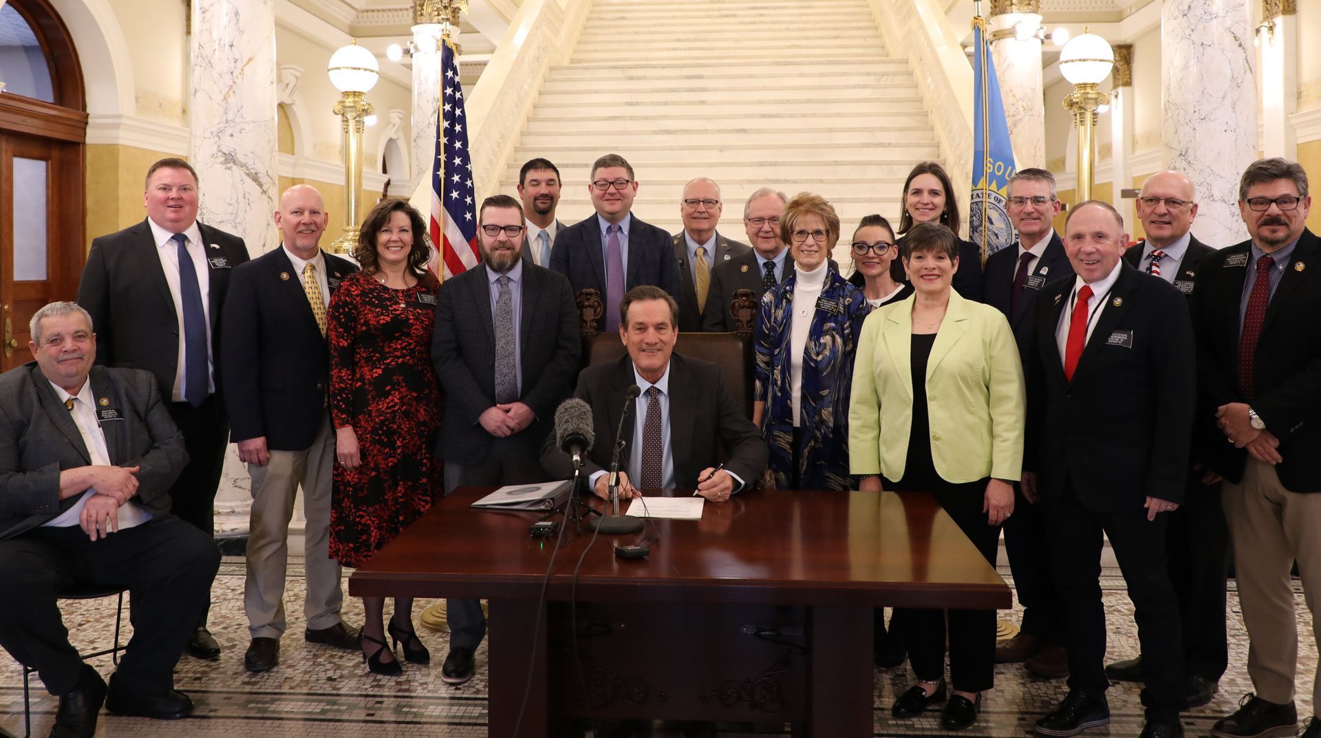 Group of officials posing in a grand hall, with one man seated at a desk in the center.