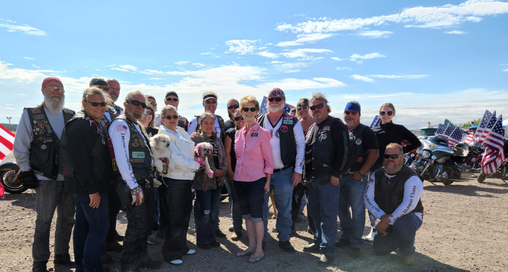 Group of people, many in leather vests, pose outdoors next to motorcycles with American flags.