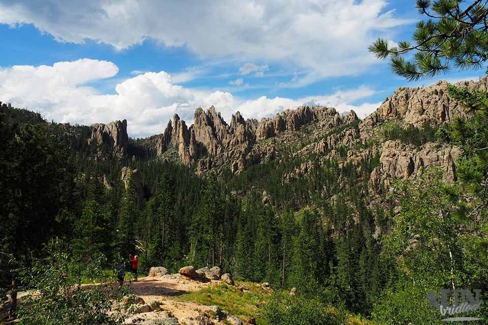 Rocky mountain peaks, lush green forest, blue sky with clouds. Two people on a hiking trail.