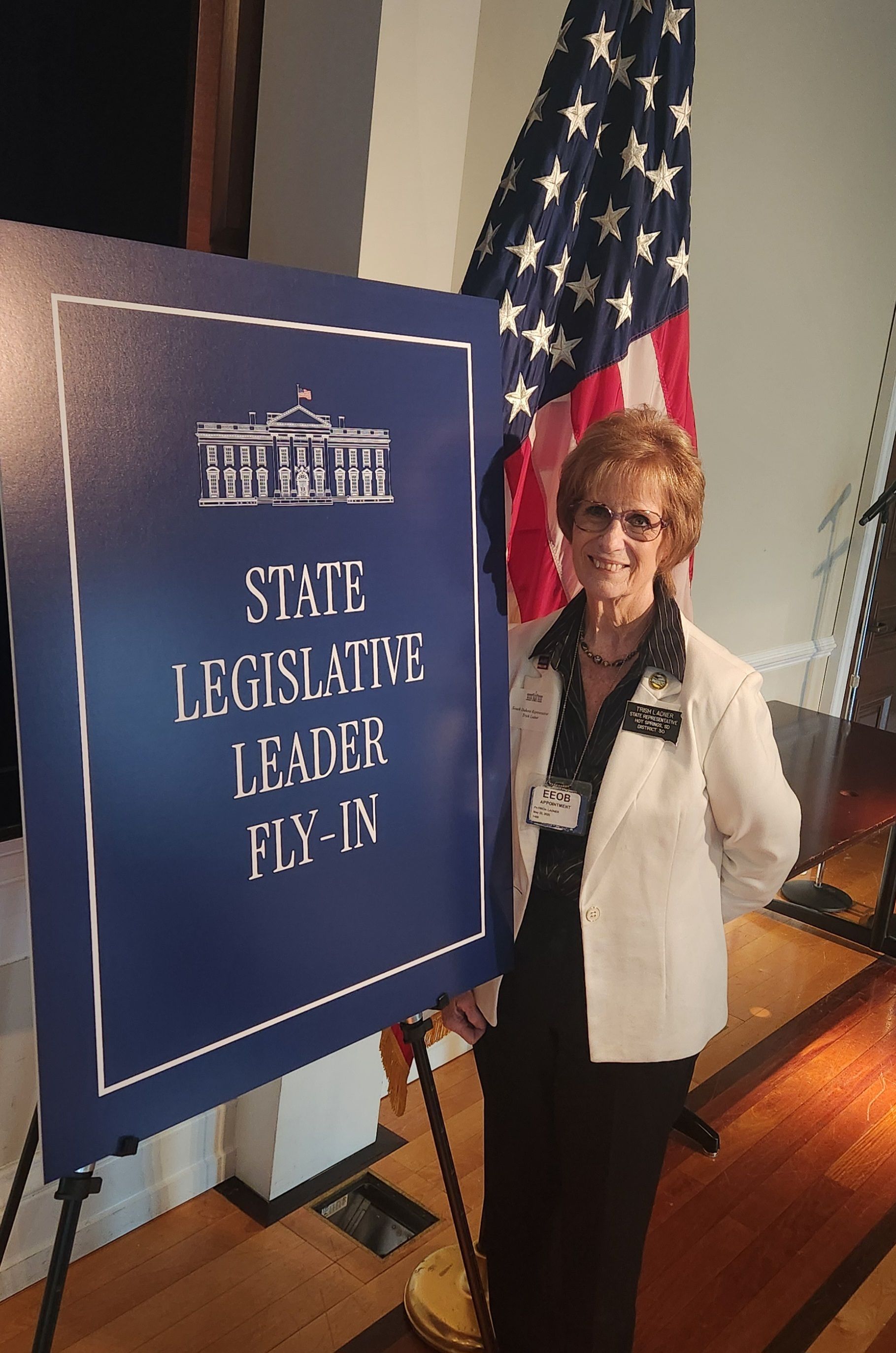 Trish standing beside a blue “State Legislative Leader” sign and U.S. flag in an indoor setting