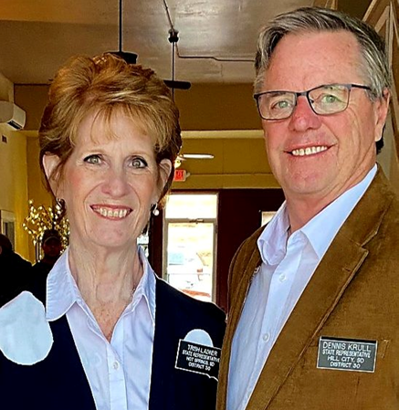 Two smiling people, a woman and a man, standing side-by-side indoors. They are wearing name tags.