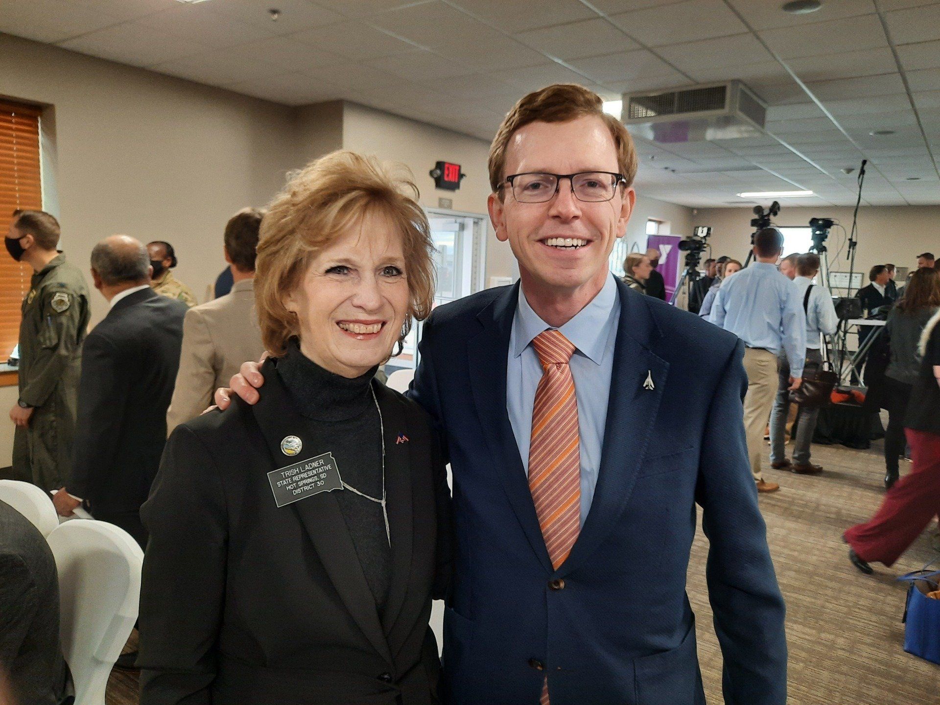 Two people posing together indoors, smiling. Man in suit, woman in black jacket. People and equipment in background.
