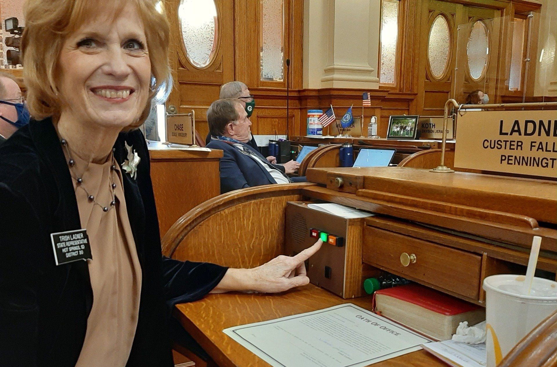 Woman in legislative chamber presses a green button, smiling. Wooden desk, papers, and other people visible.