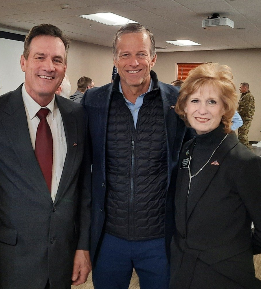 Three people smiling at the camera, inside a building. Two men in suits, one with a burgundy tie. One woman in a black blazer.