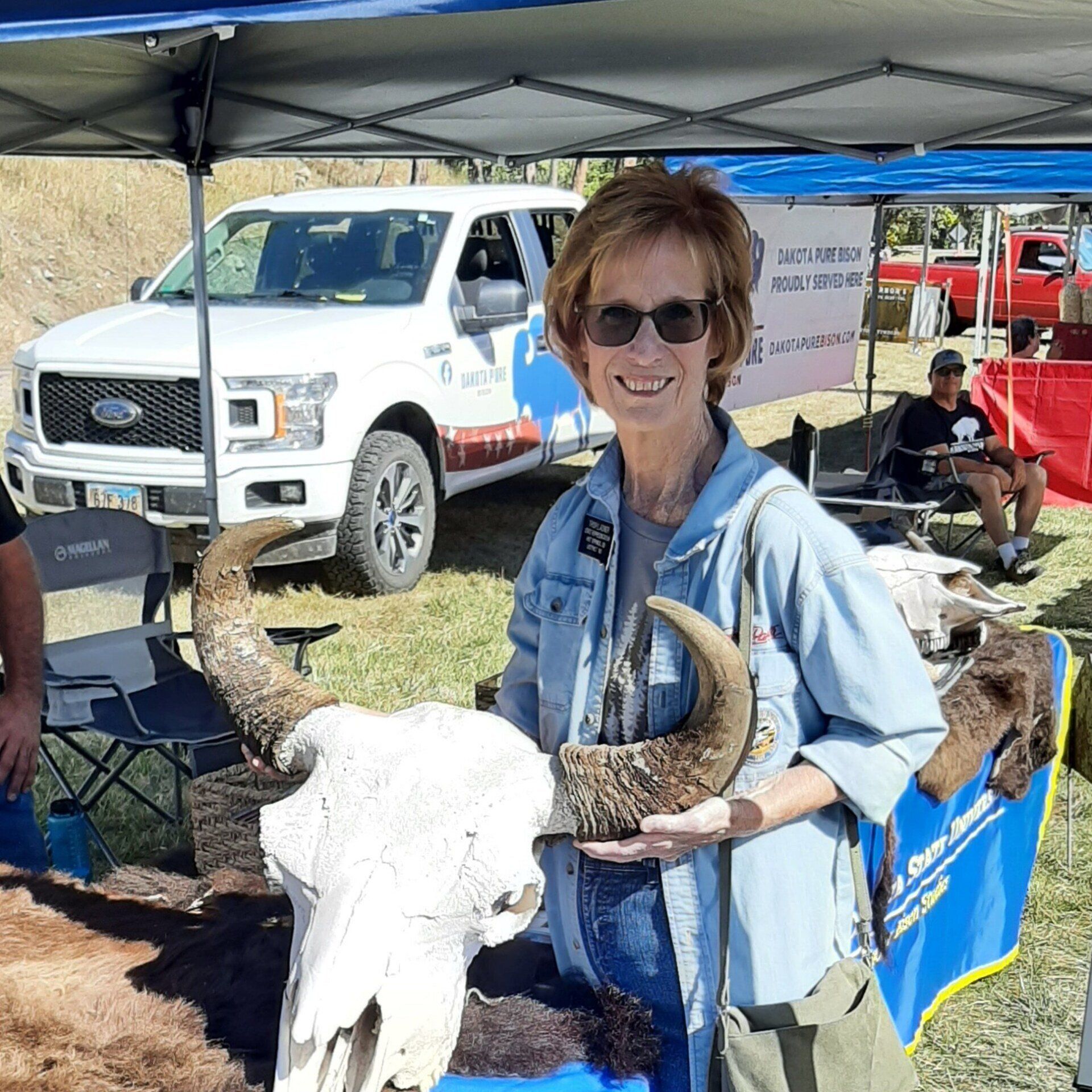 Woman holding buffalo skull and horn at an outdoor event with truck, tent, and other people in the background.