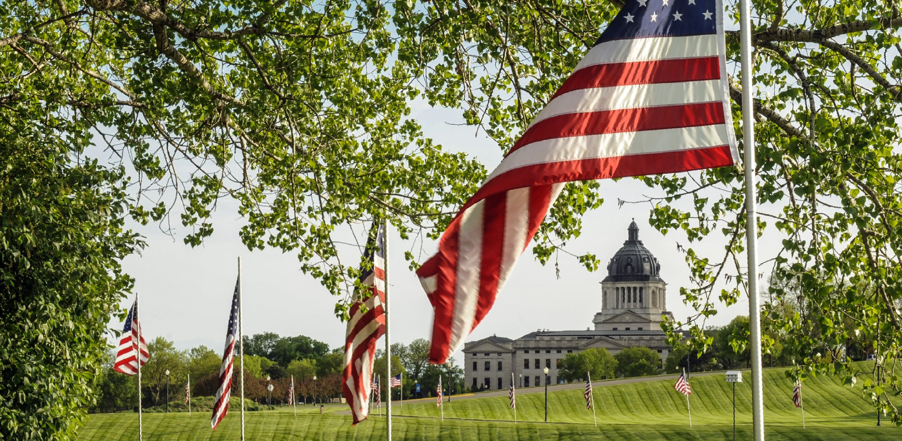 American flags fly in front of a domed building, trees frame the shot, and the sky is blue.