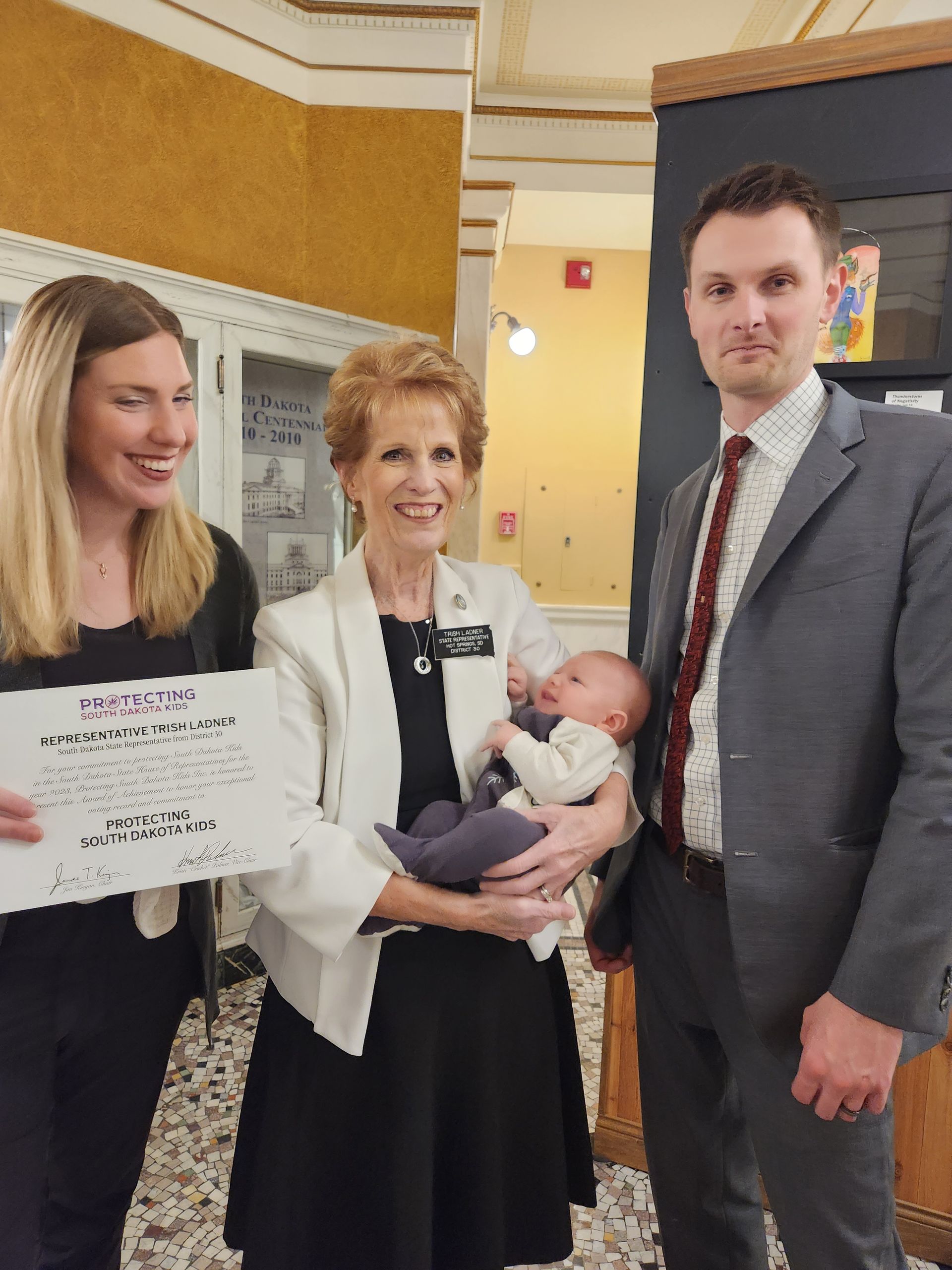 Woman holding baby, flanked by two people, holding a certificate in a hallway setting.