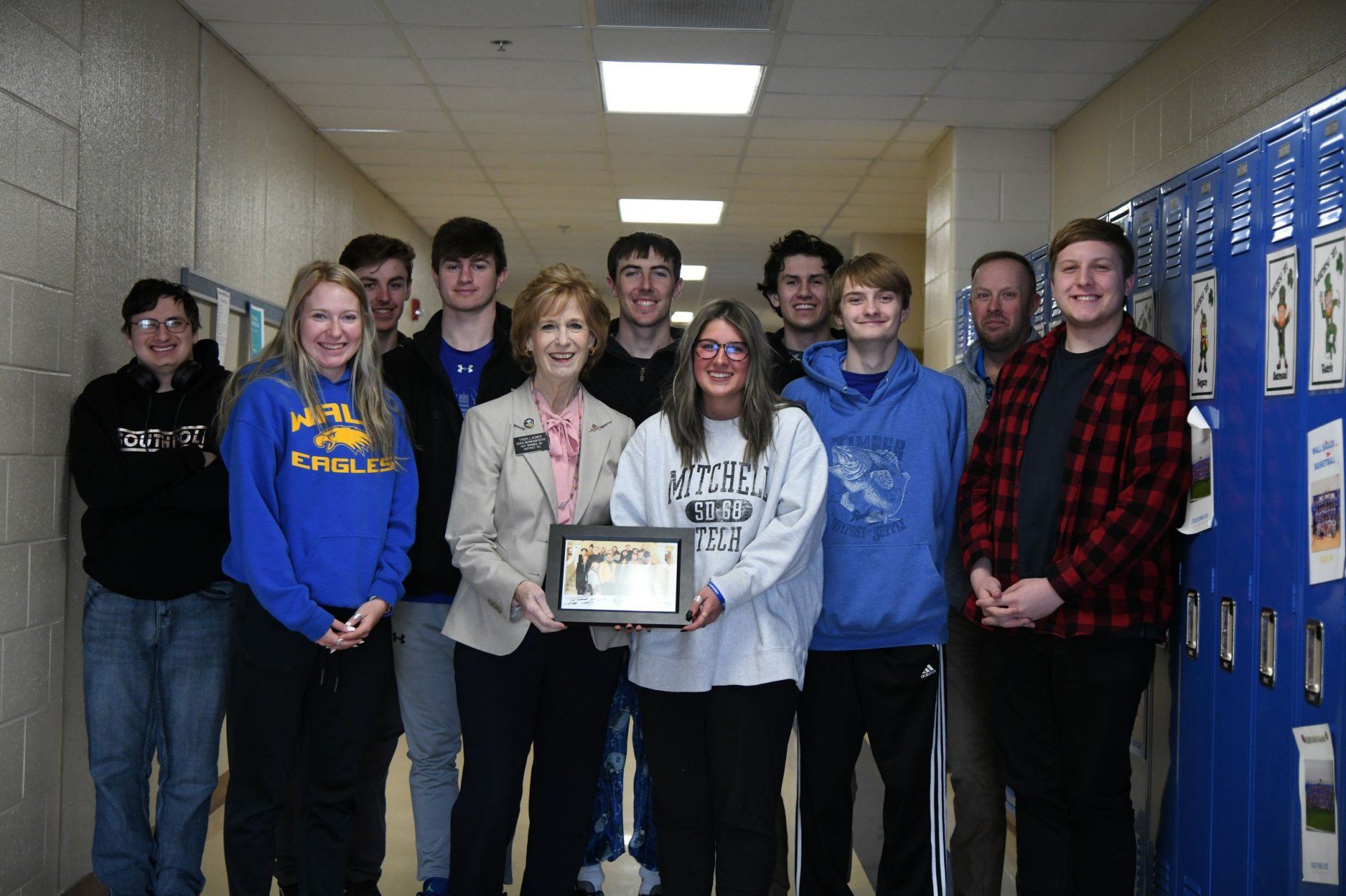 Group of people holding a framed photo in a school hallway with blue lockers.
