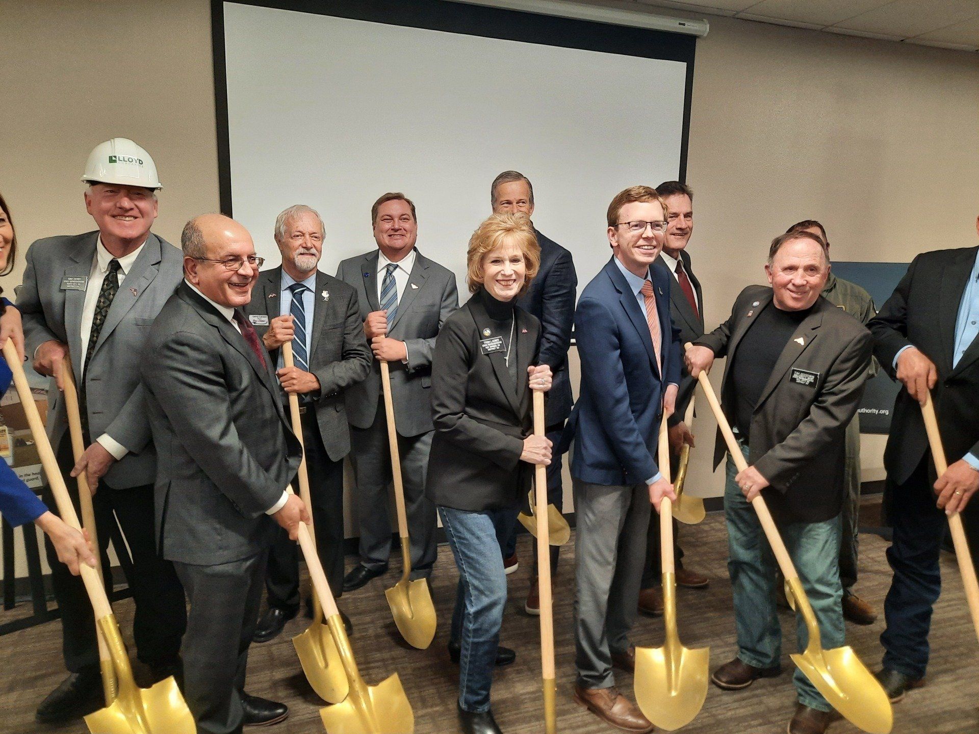 People holding shovels, ground-breaking ceremony. Indoors, neutral background.