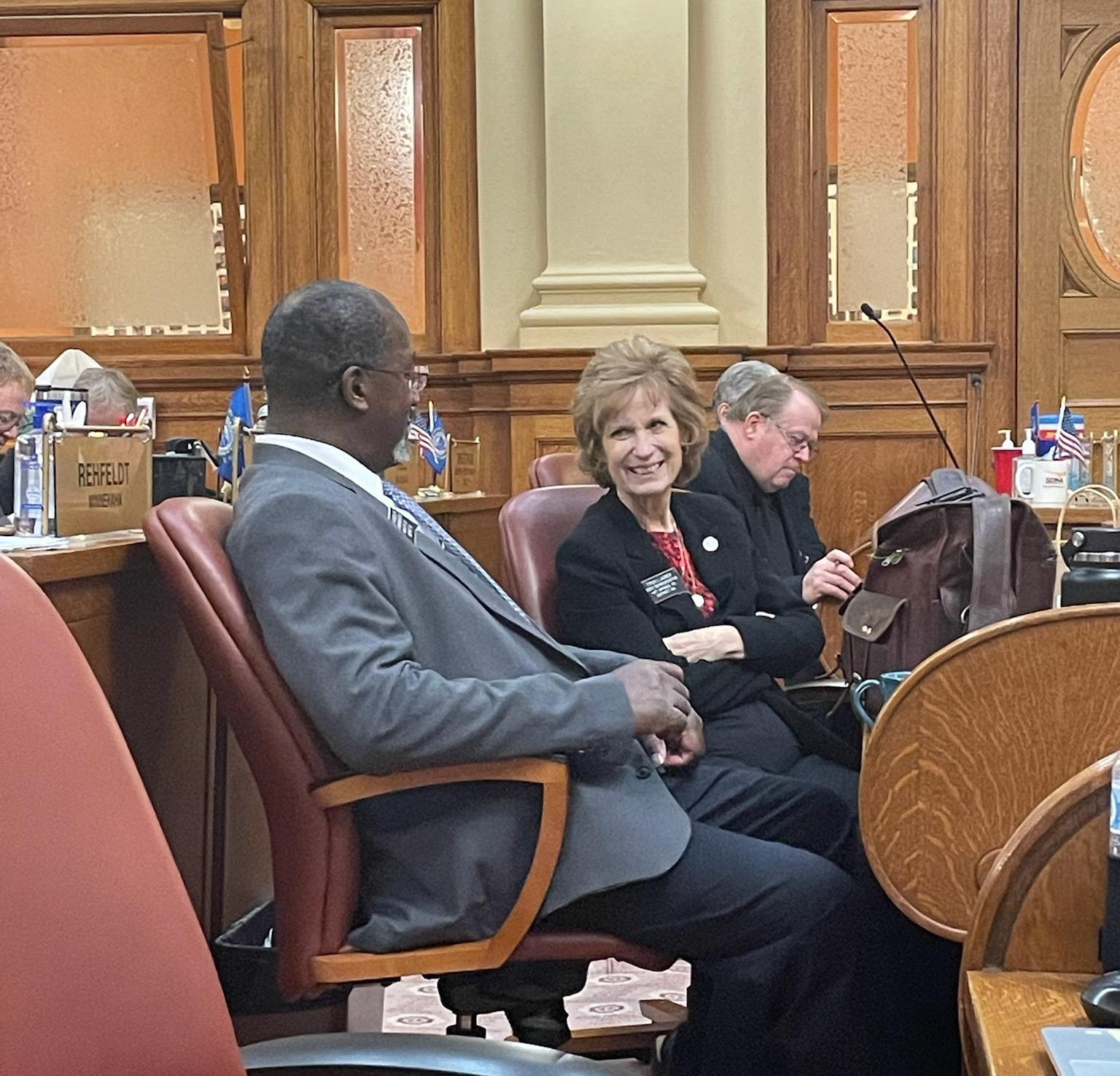 People in a meeting room, one smiles, others look forward, wooden paneling in background.