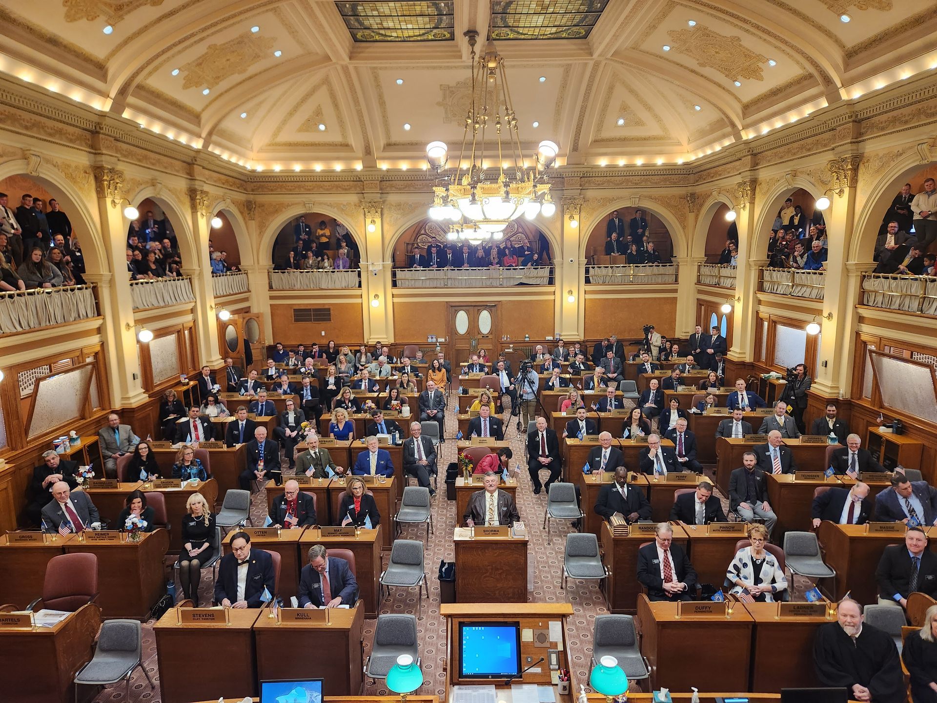 Legislative chamber with many people seated at desks, speaking. Elaborate architecture, gold accents.