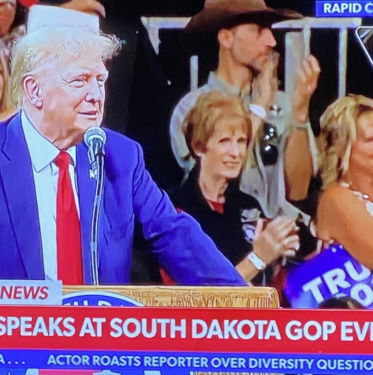 Donald Trump speaking at a South Dakota GOP event. He is wearing a blue suit and red tie, addressing a crowd.