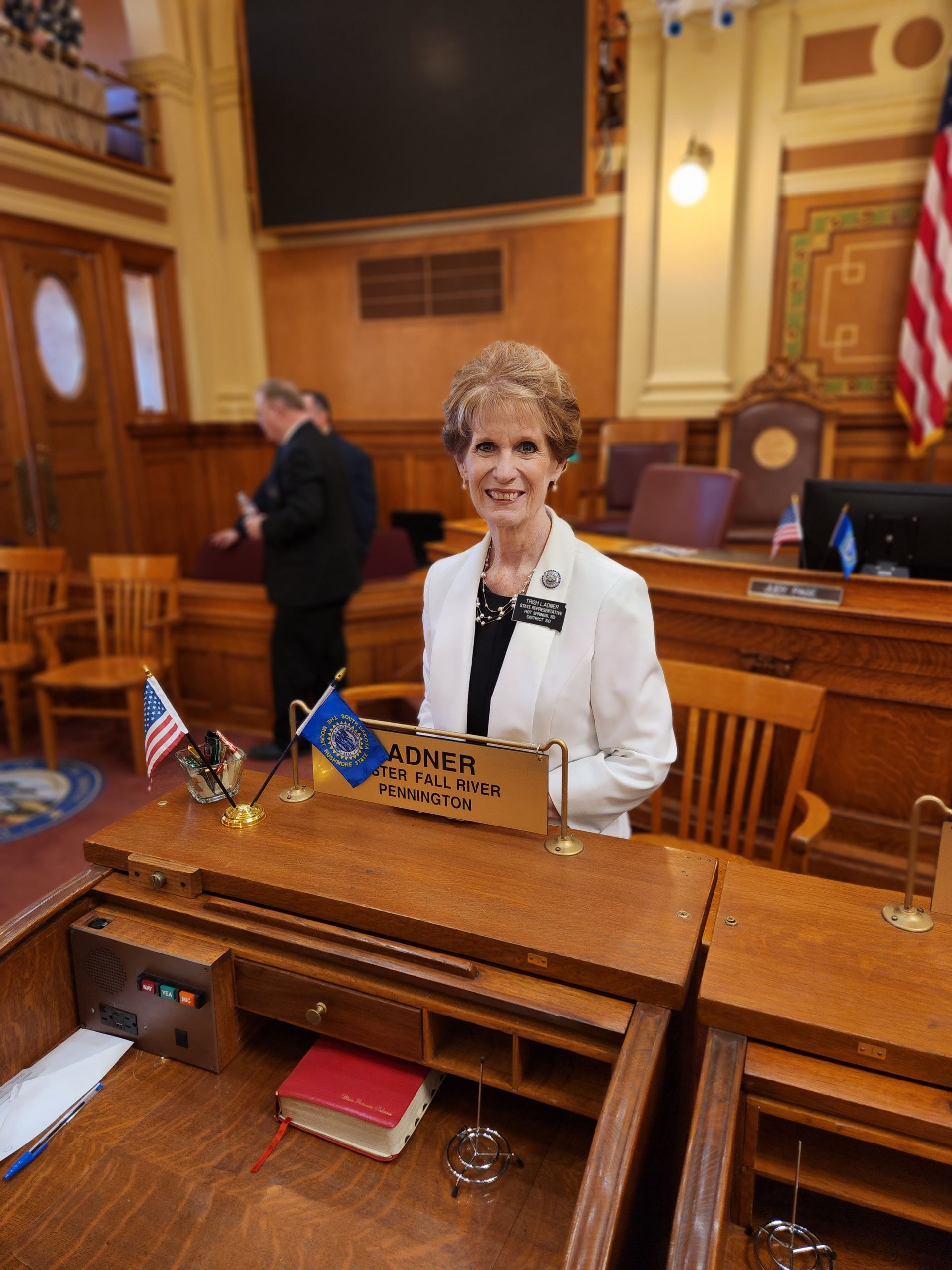 Woman in a white jacket smiles behind a desk in a legislative chamber; American flag visible.