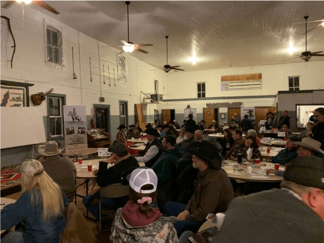 A large gathering in a hall, seated at round tables. People are listening.