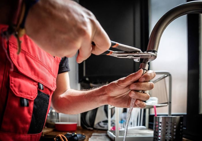 A plumbing contractor, wearing red clothing, is using adjustable pliers to fix a kitchen faucet. A plumbing contractor, wearing red clothing, is using adjustable pliers to fix a kitchen faucet.