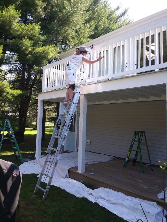 Person on a ladder painting a white porch railing on a house exterior