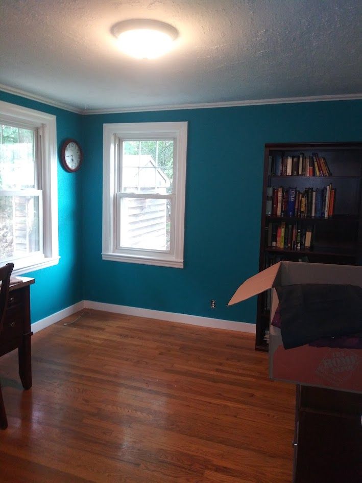 Empty room with teal walls, hardwood floors, two windows, ceiling light, and a bookshelf with a covered chair.