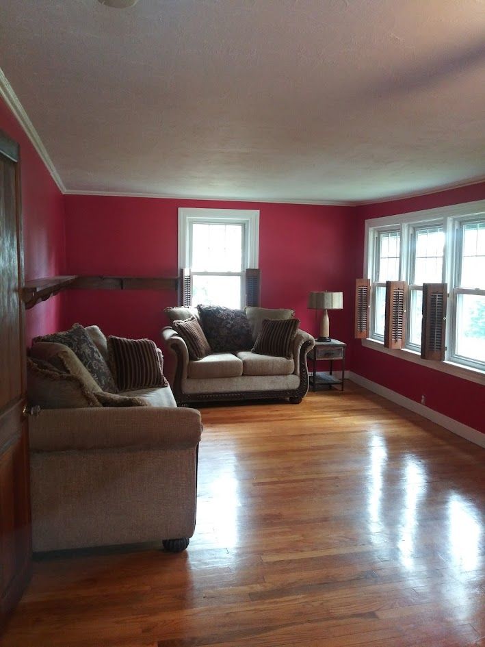 Bright living room with red walls, hardwood floor, beige couches, and large windows.