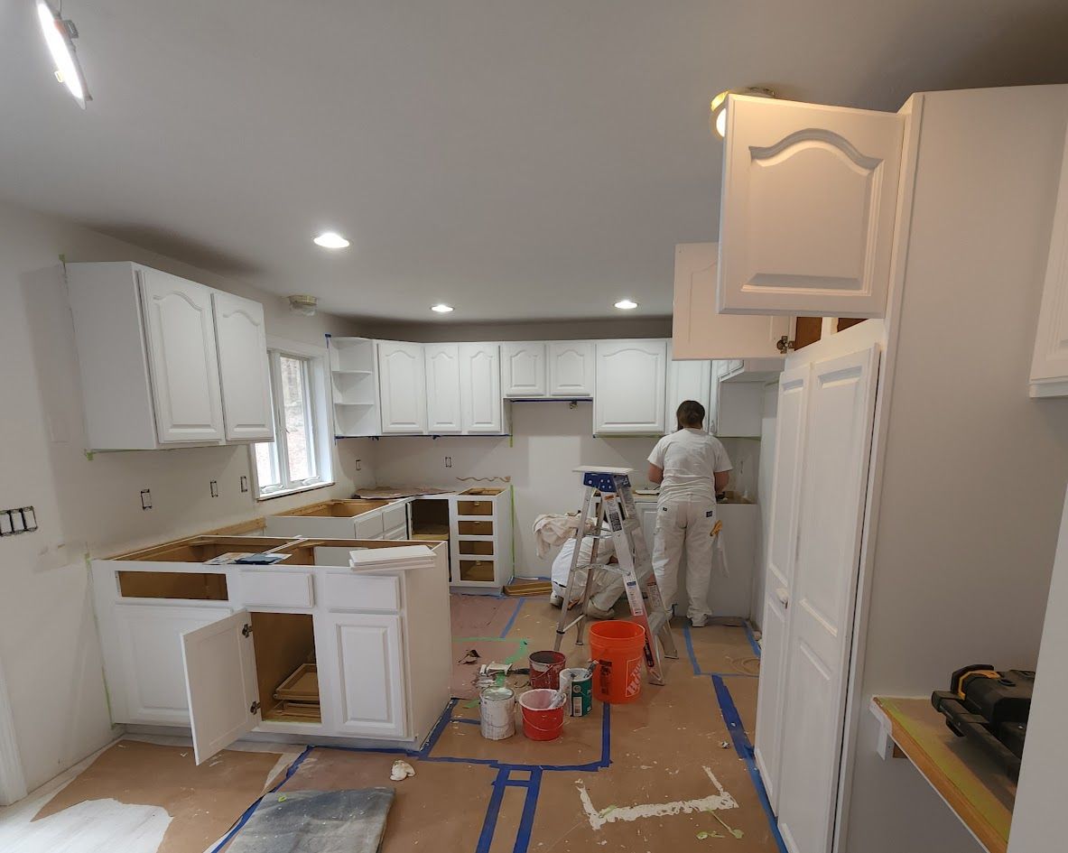Kitchen under renovation with white cabinets, tools, and a worker painting near the back wall