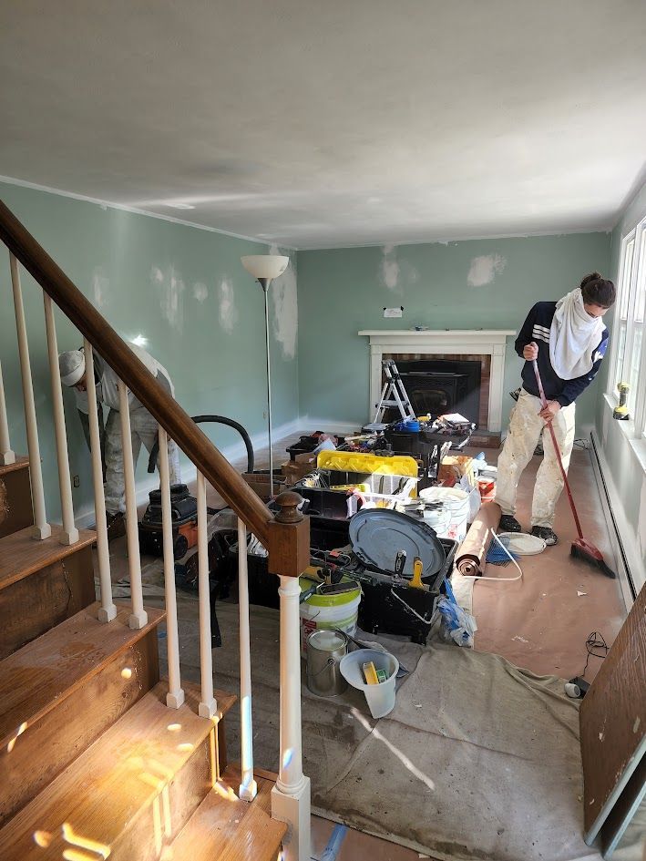Messy hallway renovation with tools and debris; worker stands at right beside staircase.