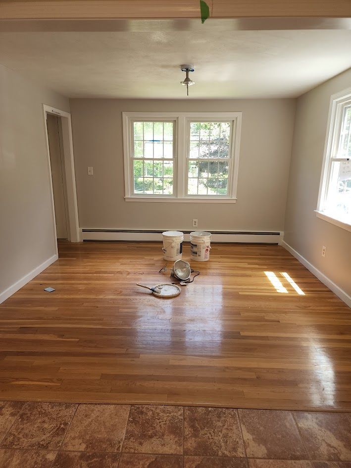 Empty living room with hardwood floors being refinished, paint bucket and tools in the center.