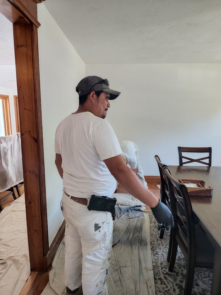 Person in white work clothes and gloves cleaning a dining table in a bright home interior.