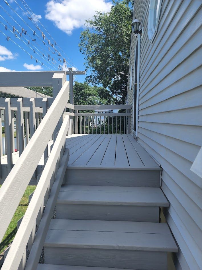 White outdoor stairs leading to a small deck beside a house, with blue sky and trees overhead.