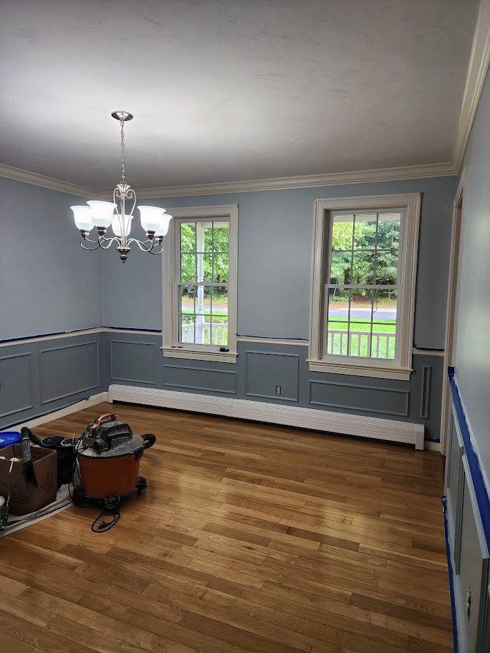 Empty room with blue-gray wainscoted walls, hardwood floor, chandelier, and two windows.