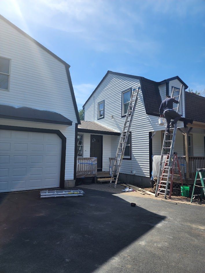 Driveway view of a house exterior with garage, gray siding, and a ladder under a blue sky.