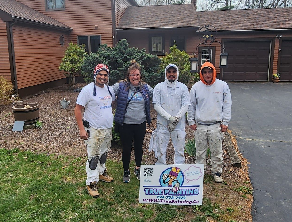 Four people standing outdoors by a sign in front of a house and garage, smiling.