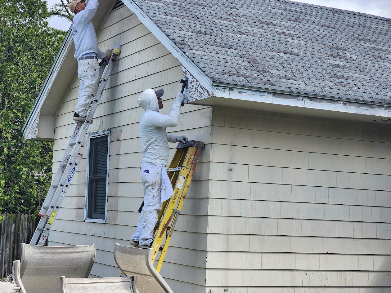 Two painters in white coveralls painting the exterior siding of a beige house from ladders.