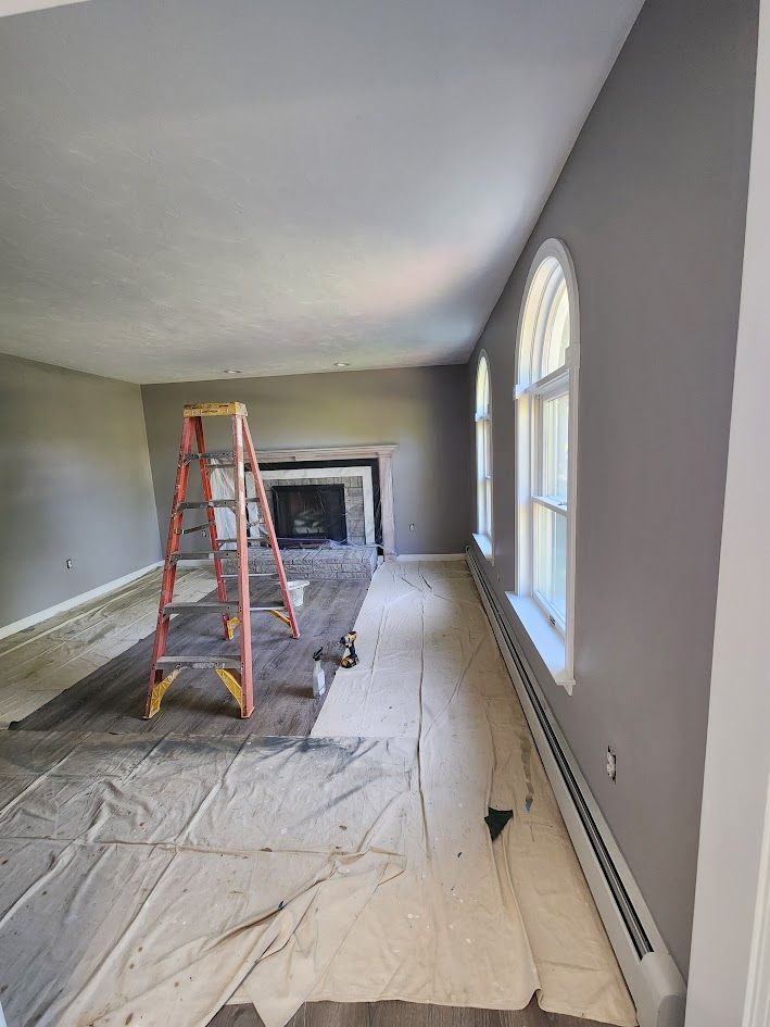 Renovation in progress in a long room with a ladder, fireplace, tarps, and arched windows.