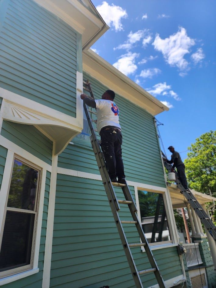 Workers painting exterior siding on a house from ladders under a blue sky.