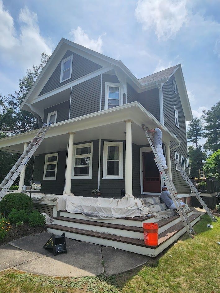Two-story house with dark siding and white trim, undergoing exterior renovation with ladders and scaffolding.